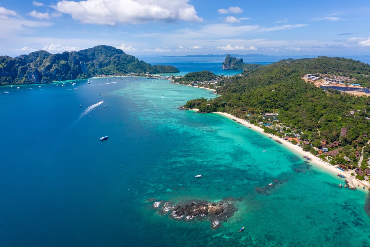 Alt Text: Aerial view of a tropical beachfront resort and a tourist boat near the shores of Phi Phi Island in Krabi, Thailand.